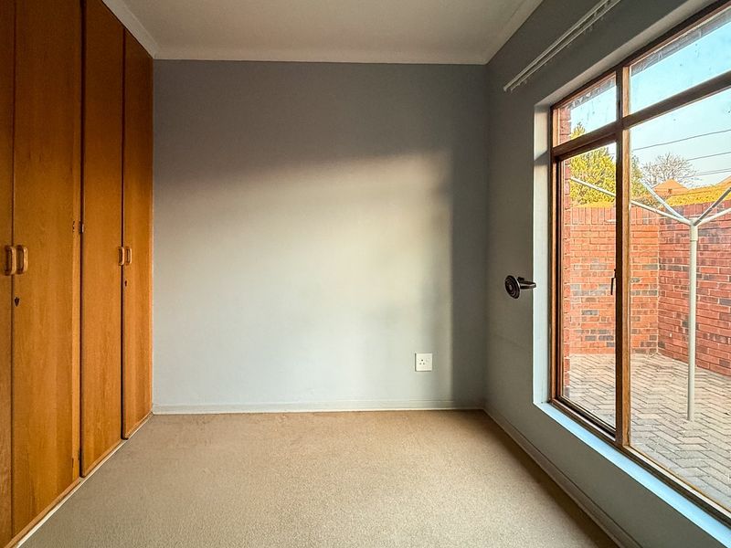 An empty bedroom featuring built-in wooden cupboards, carpeted flooring, and a large window overlooking a private paved courtyard.