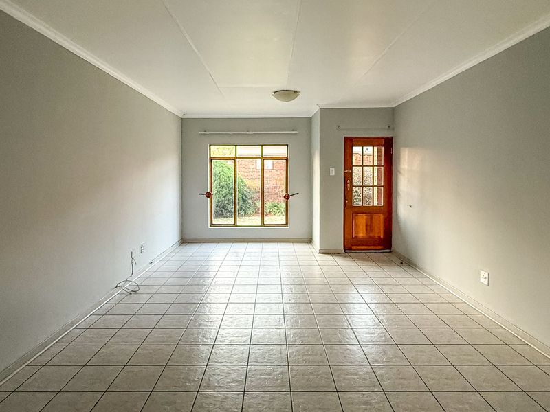 A bright, empty living room featuring neutral walls, ceramic tile flooring, and a classic timber-framed glass door leading to the exterior.