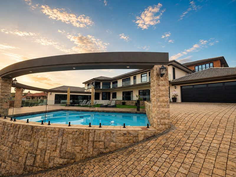 A luxury exterior view featuring a swimming pool with glass balustrades, stone-clad retaining walls, and a paved driveway leading to a garage.