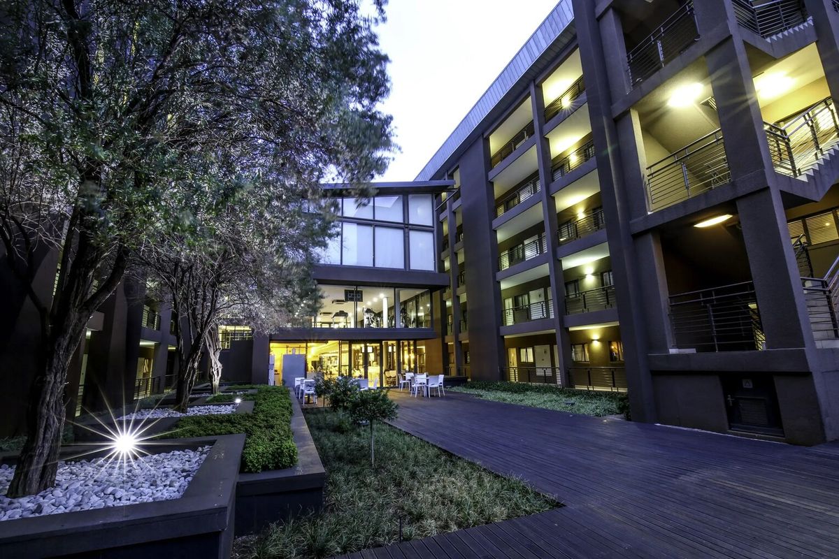 A well-maintained apartment complex courtyard featuring timber decking, landscaped garden beds, and multi-story residential balconies illuminated by exterior lighting.