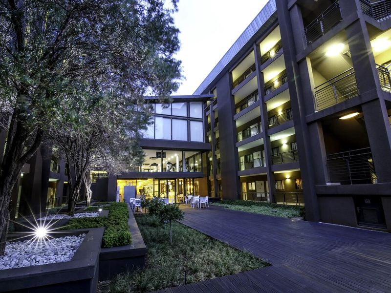 A well-maintained apartment complex courtyard featuring timber decking, landscaped garden beds, and multi-story residential balconies illuminated by exterior lighting.