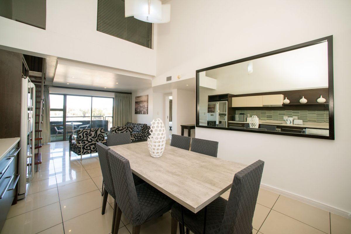 Spacious open-plan living and dining area featuring high ceilings, tiled floors, and seamless flow to an outdoor balcony, with an adjacent kitchen visible.