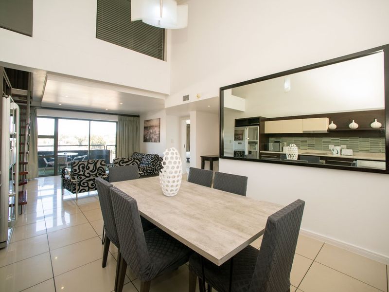 Spacious open-plan living and dining area featuring high ceilings, tiled floors, and seamless flow to an outdoor balcony, with an adjacent kitchen visible.