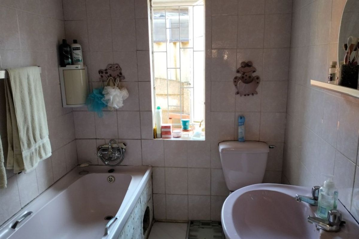 A functional bathroom featuring a bathtub, pedestal basin, and toilet with full-height wall tiling and a window with security bars.