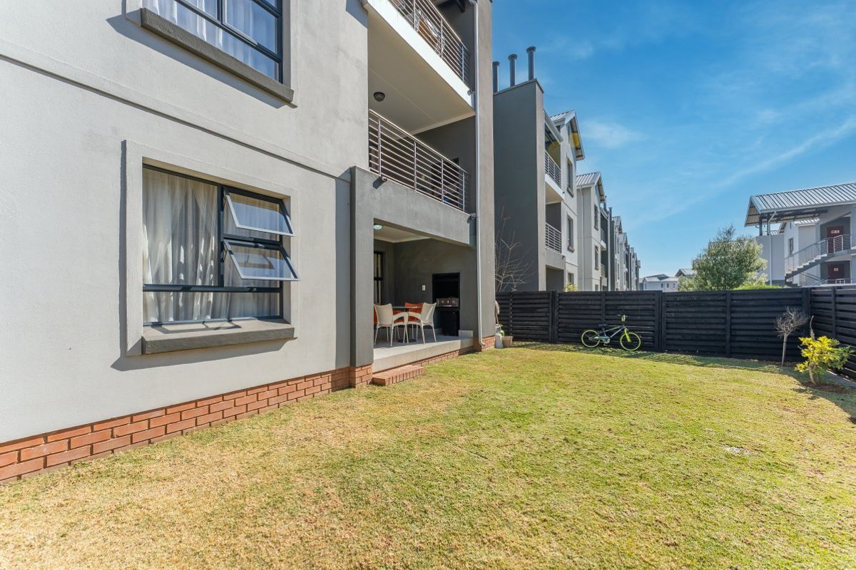 Rear view of the apartment building featuring a private garden, a covered patio with a built-in braai, and upper-level balconies with metal railings.