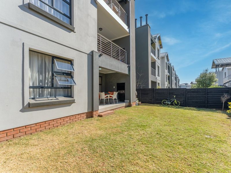Rear view of the apartment building featuring a private garden, a covered patio with a built-in braai, and upper-level balconies with metal railings.