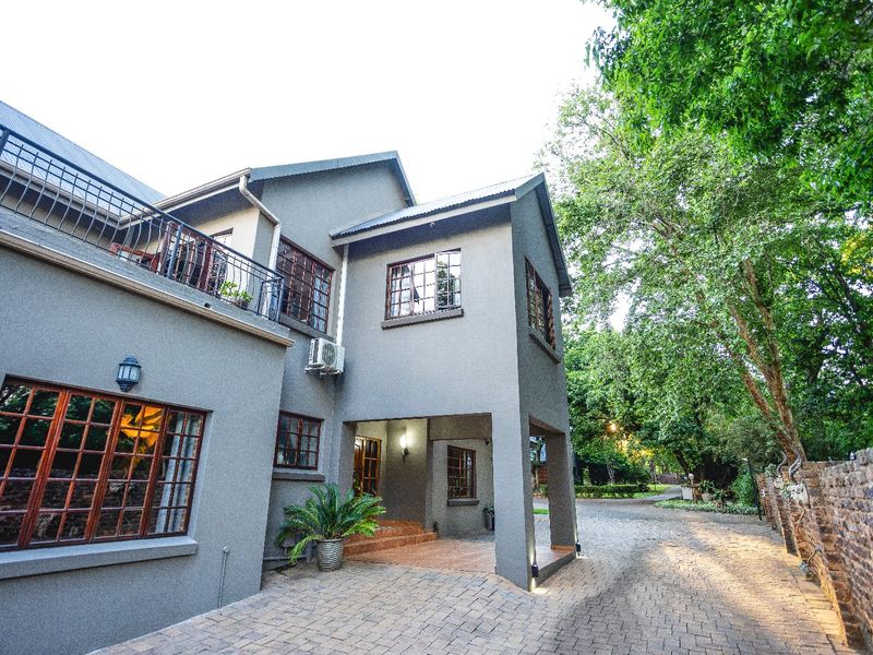 A well-maintained double-story house featuring a paved driveway, covered entrance, and a balcony with metal railings, set within a lush, tree-lined property.