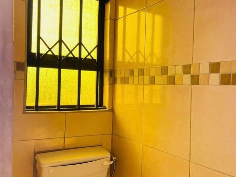 A compact bathroom featuring neutral ceramic wall tiles, a functional toilet cistern, and a window fitted with security bars.