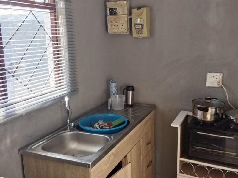 A compact kitchen area featuring a stainless steel sink, basic cabinetry, and exposed electrical infrastructure, requiring cosmetic updates and repairs to finishes.