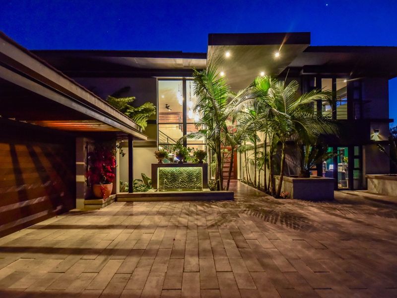 Modern architectural front facade at dusk, featuring a paved driveway, covered entrance, integrated water feature, and extensive glass detailing.