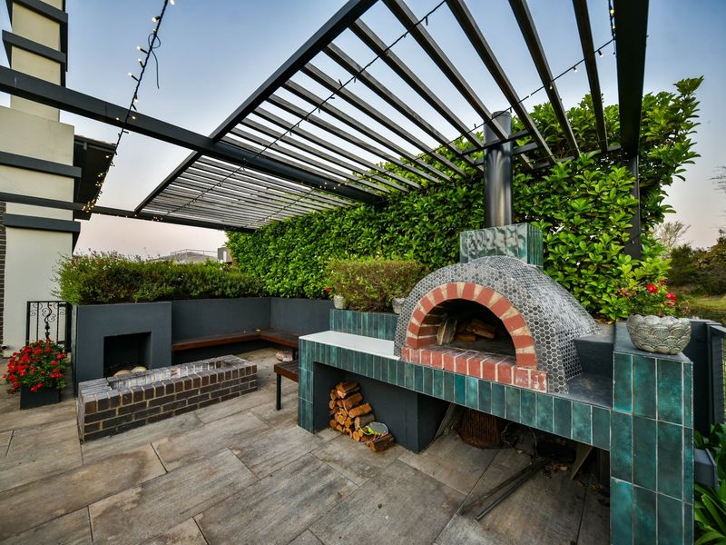 Outdoor entertainment patio featuring a built-in pizza oven, fireplace, and pergola, set against a landscaped garden backdrop.