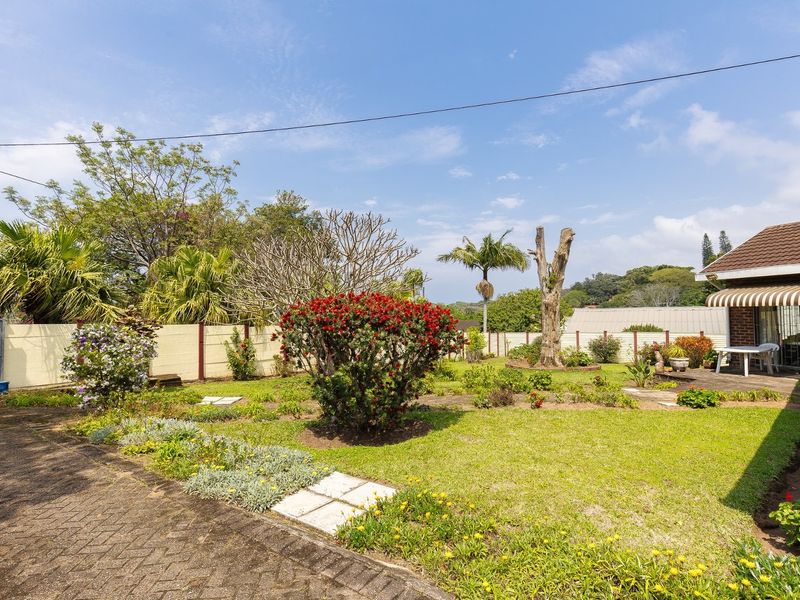 A spacious rear garden featuring a paved driveway, established landscaping, a precast boundary wall, and a covered patio area attached to the main house.