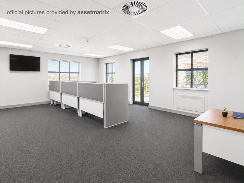 A bright, modern open-plan office space featuring carpeted floors, suspended ceilings with recessed lighting, and direct access to an outdoor balcony area.