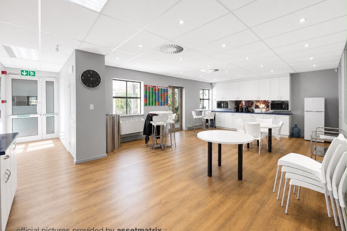 A bright, modern commercial kitchen and breakroom area featuring white cabinetry, wood-look laminate flooring, and a suspended ceiling with recessed lighting.
