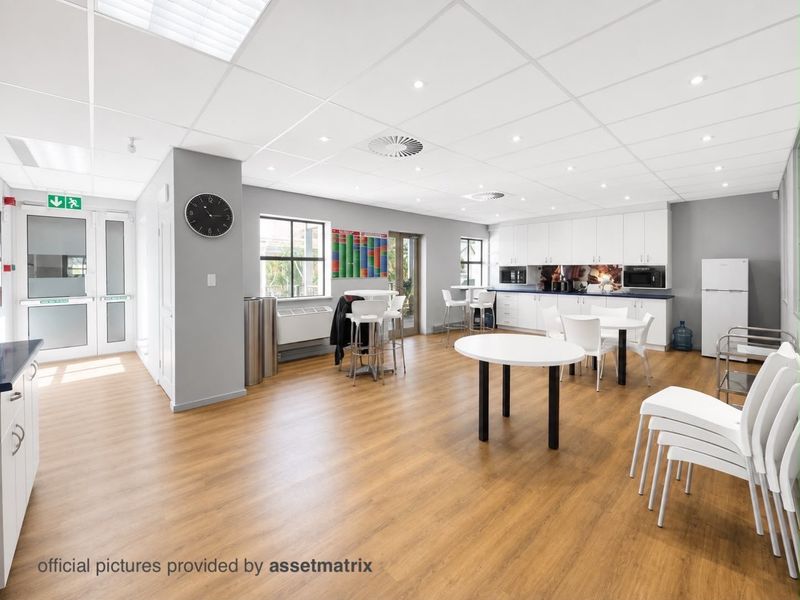 A bright, modern commercial kitchen and breakroom area featuring white cabinetry, wood-look laminate flooring, and a suspended ceiling with recessed lighting.