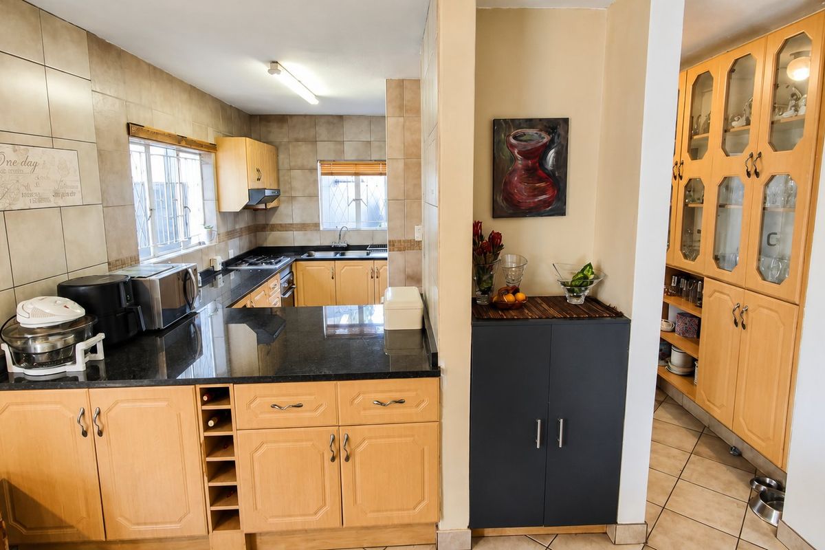 A functional kitchen featuring granite countertops, light wood cabinetry, glass-fronted display units, and neutral ceramic tiling throughout the workspace and adjacent storage area.