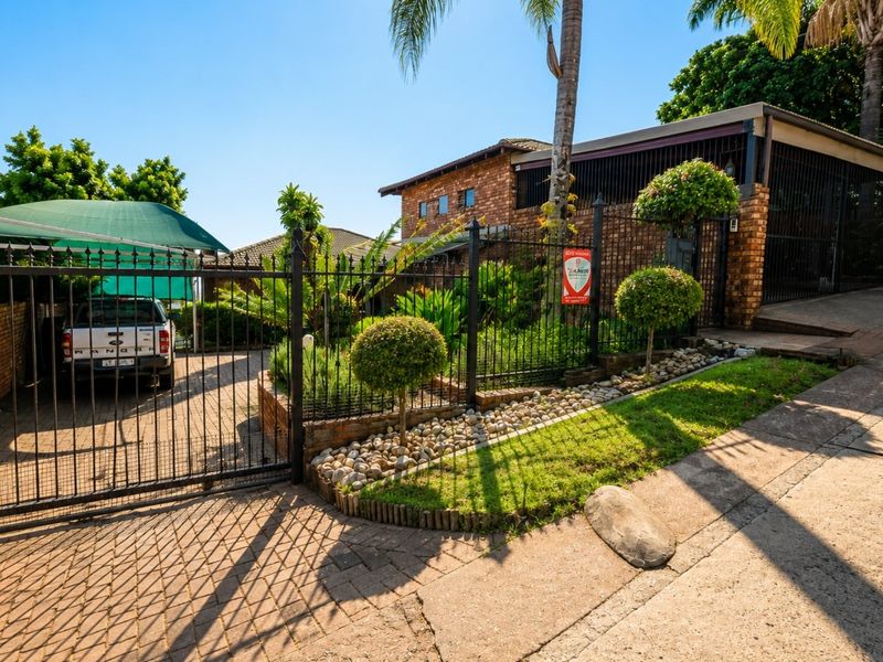 Front exterior view of a brick house featuring a paved driveway, secure wrought iron fencing, a carport, and a landscaped garden area.