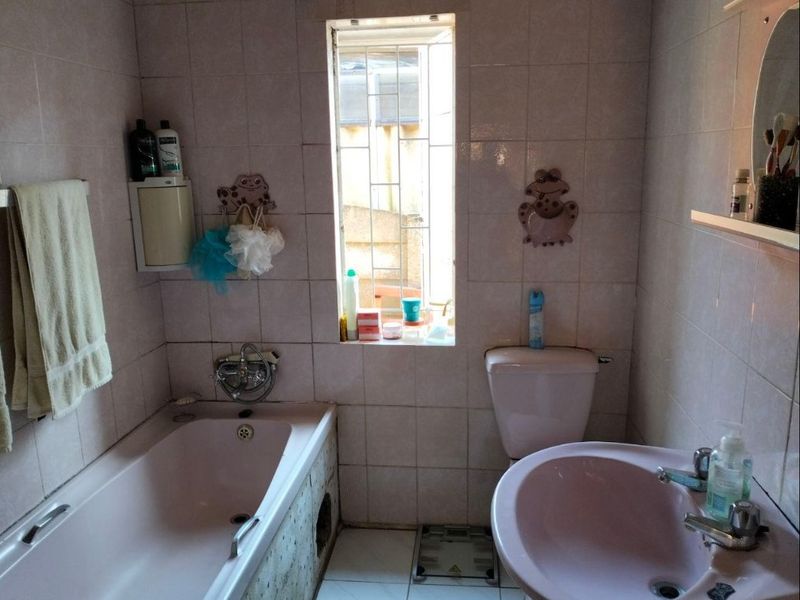 A dated bathroom featuring a pink bathtub and sink suite with full-height wall tiling and a window with security bars.