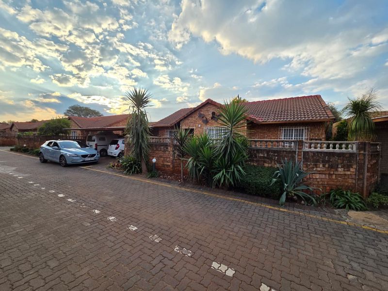 Front exterior view of a facebrick townhouse featuring a tiled roof, carport, and secure boundary wall with established garden elements.