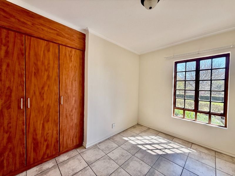 A bright, empty bedroom featuring tiled flooring, a built-in wooden wardrobe, and a grid-pattern window overlooking the garden.