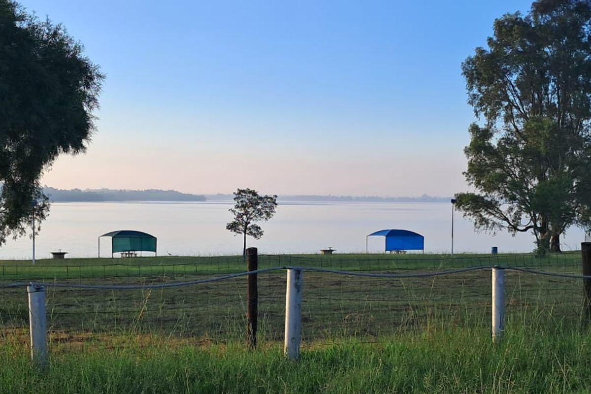 A scenic view of a waterfront area featuring open green space, picnic shelters, and mature trees, enclosed by a wire fence.