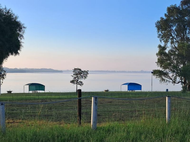 A scenic view of a waterfront area featuring open green space, picnic shelters, and mature trees, enclosed by a wire fence.