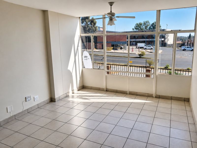A bright, empty living area featuring tiled flooring, a ceiling fan, and large windows providing natural light and street views.