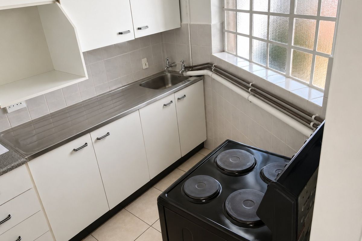 A compact kitchen featuring white cabinetry, a stainless steel sink, tiled splashbacks, and an electric stove, with natural light from a grid window.