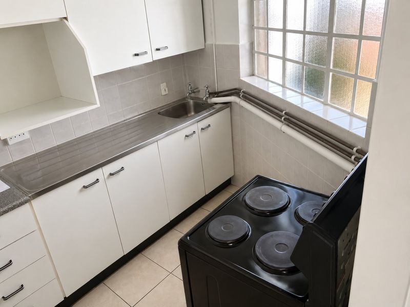 A compact kitchen featuring white cabinetry, a stainless steel sink, tiled splashbacks, and an electric stove, with natural light from a grid window.