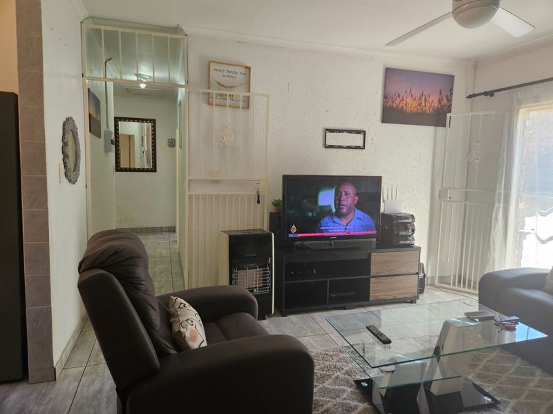 A living room featuring tiled flooring, a ceiling fan, and a security gate, with visible wall wear near the hallway entrance.