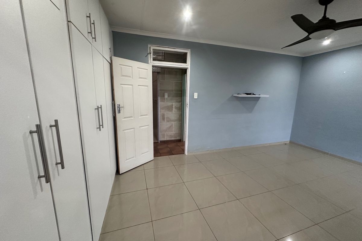 A spacious, empty bedroom featuring neutral tiled flooring, extensive built-in cupboard storage, and a ceiling fan, with direct access to an adjacent tiled area.