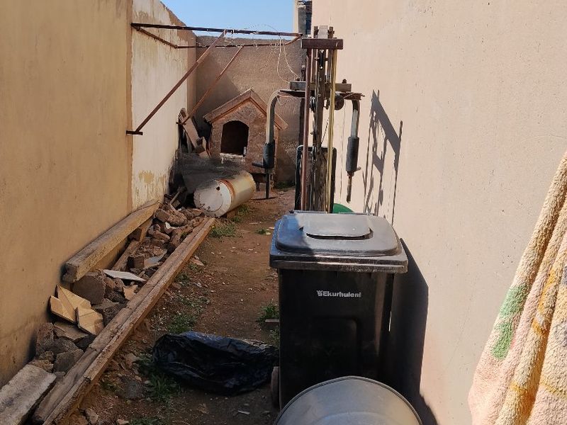 A narrow side or rear exterior passage featuring high boundary walls and an unpaved ground surface requiring maintenance and clearing.