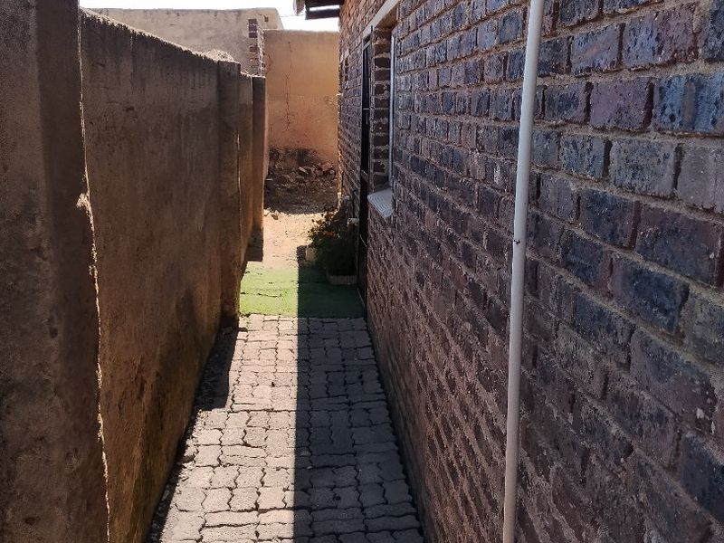 A narrow side passage featuring exposed brickwork, a paved walkway, and a high boundary wall, providing access along the side of the house.