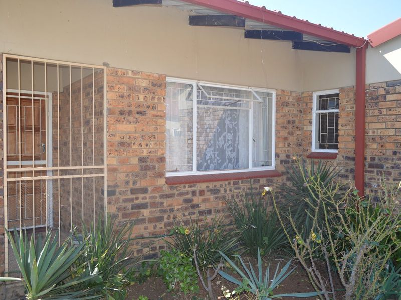Front facade of a face brick townhouse featuring security gates, metal window frames, and a corrugated metal roof with exposed rafters.