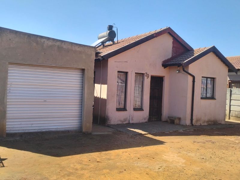 Front elevation of a single-story house featuring a roller garage door, solar water heater, and tiled roof with an unpaved driveway area.