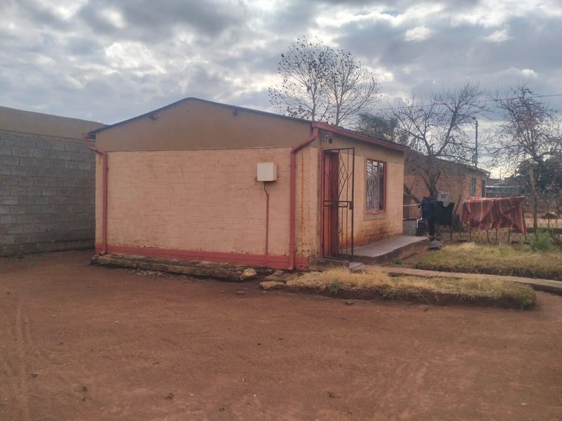 Rear view of a single-story residential dwelling featuring masonry construction, metal security gate, and basic guttering on a dirt plot.