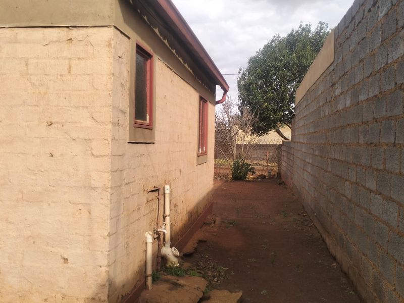 A side exterior view of the property showing exposed brick walls, external plumbing, and a concrete block boundary wall with unpaved ground.