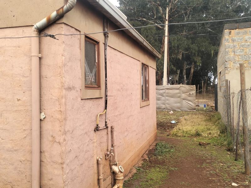 Rear exterior view of the property showing weathered plaster walls, external plumbing, and a damaged chain link fence boundary.