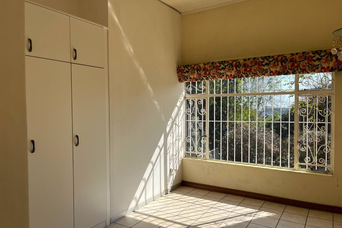 A bright, empty bedroom featuring built-in cupboards, tiled flooring, and large windows fitted with security bars.