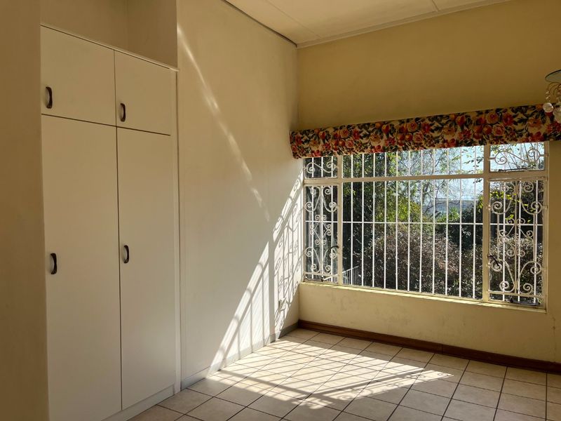 A bright, empty bedroom featuring built-in cupboards, tiled flooring, and large windows fitted with security bars.
