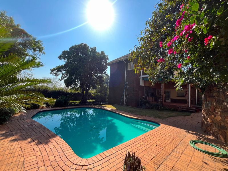 A well-maintained outdoor pool area featuring brick paving, a covered patio, and established garden surroundings.