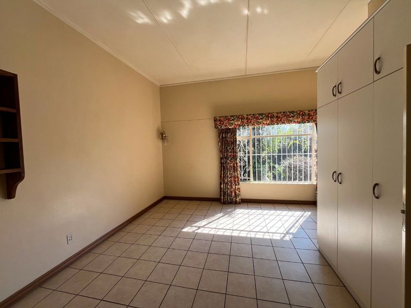 A spacious, empty bedroom featuring neutral tiled flooring, built-in cupboards, and a window fitted with security bars.