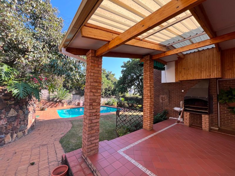 Covered outdoor patio featuring a built-in braai, brick pillars, and timber pergola, overlooking a private swimming pool and garden area.