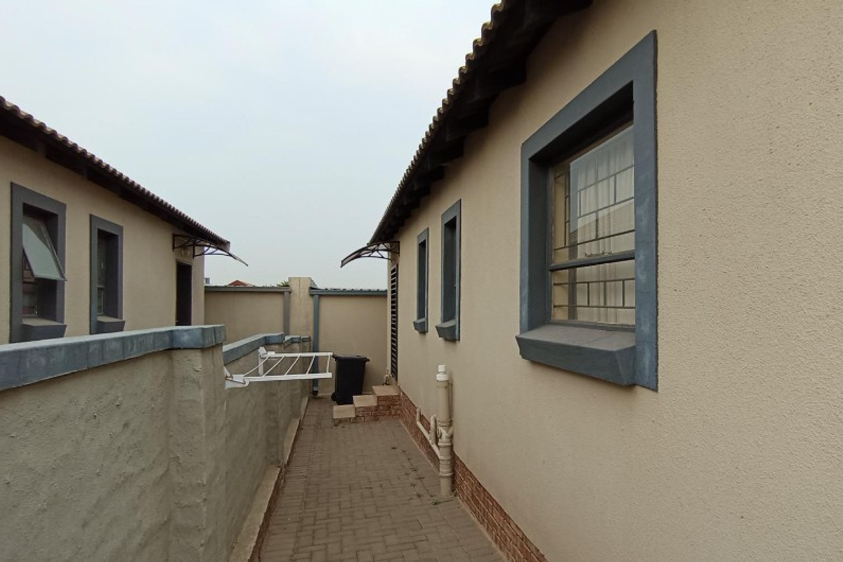 A side exterior walkway between two residential structures featuring paved flooring, security bars on windows, and perimeter boundary walls.