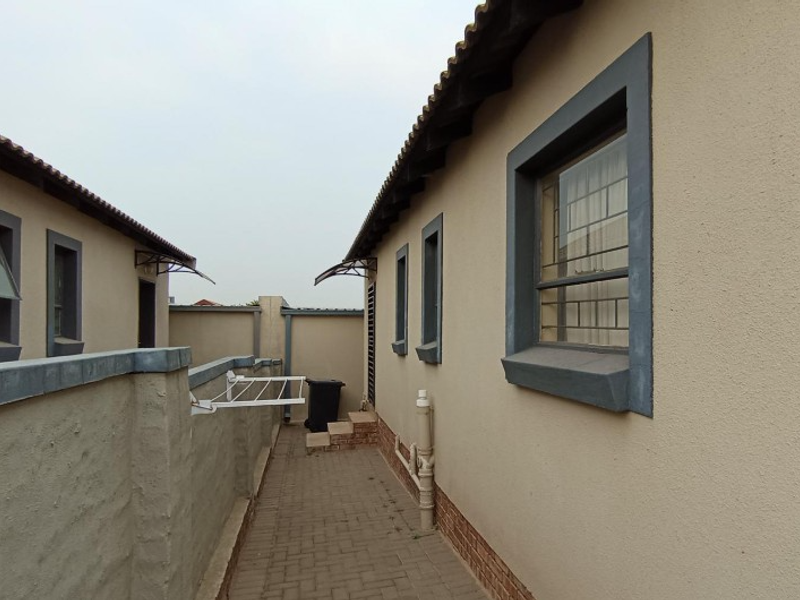 A side exterior walkway between two residential structures featuring paved flooring, security bars on windows, and perimeter boundary walls.