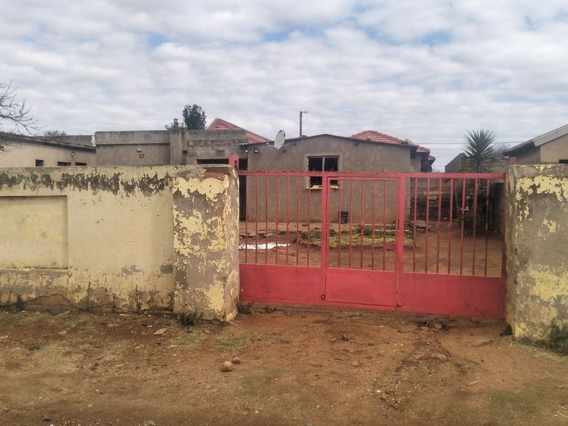 Front view of a residential property featuring a perimeter wall and a red metal driveway gate, showing signs of exterior wear.