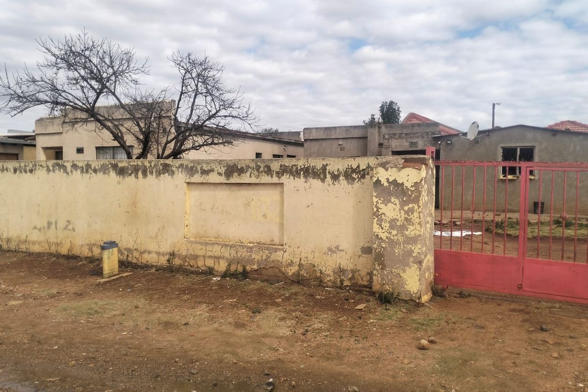Front view of the property featuring a weathered perimeter wall and a metal entrance gate, showing significant signs of exterior wear.