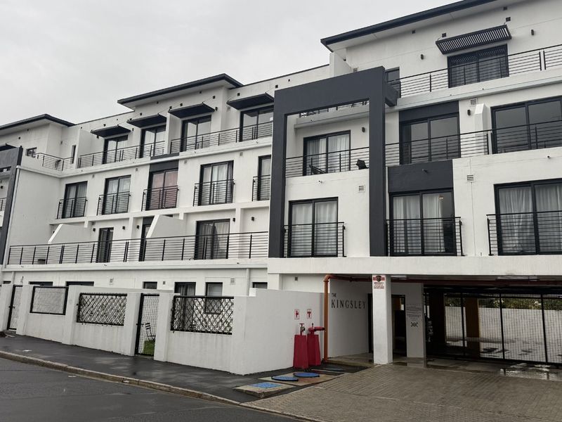 A modern multi-story apartment complex featuring white and dark grey facade, private balconies with metal railings, and a secure gated entrance.