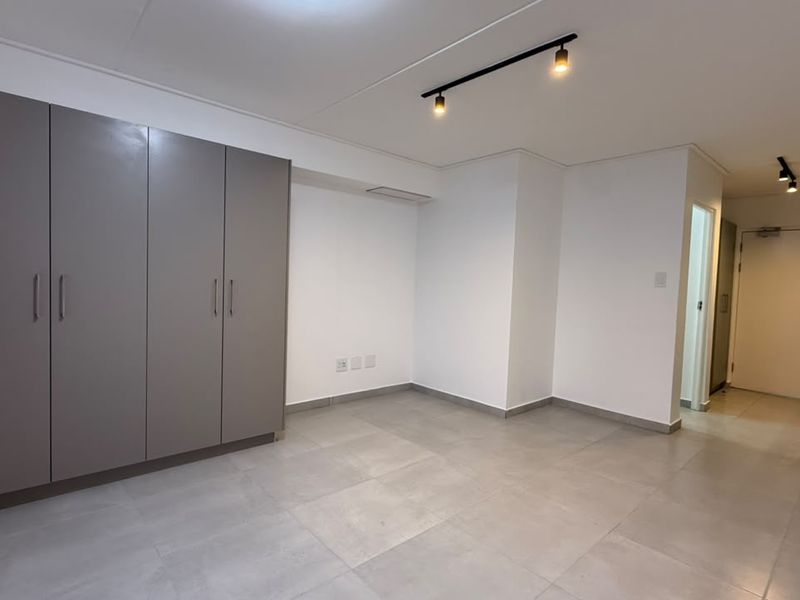 Modern open-plan living area featuring neutral tiled flooring, built-in storage cupboards, and integrated kitchen fixtures with contemporary track lighting.