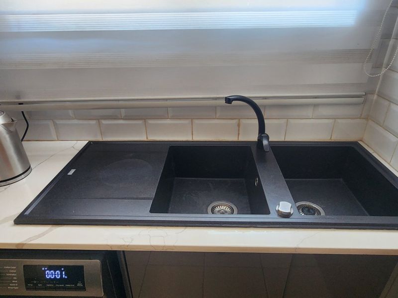 A modern kitchen area featuring a double bowl sink with a black mixer tap, stone countertop, and white subway tile backsplash.